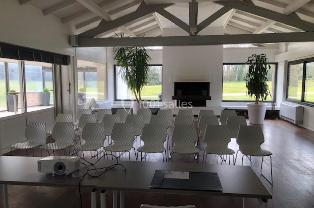 Salle lumineuse avec des chaises blanches alignées, un bureau avec un projecteur au premier plan et de grandes fenêtres.