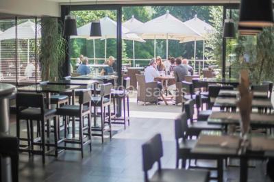 Salle lumineuse avec baies vitrées, tables rondes dressées avec nappes blanches et chaises modernes.