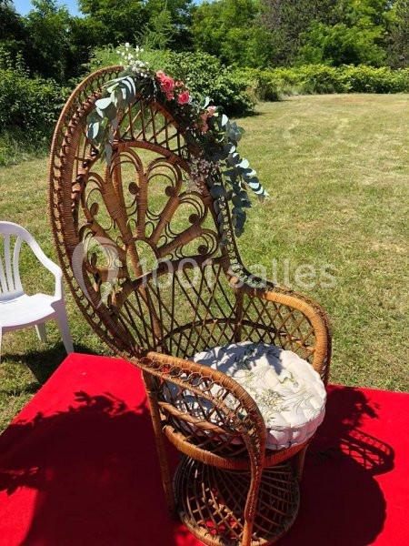 Fauteuil en rotin décoré de fleurs et feuillages, placé sur un tapis rouge dans un jardin ensoleillé.