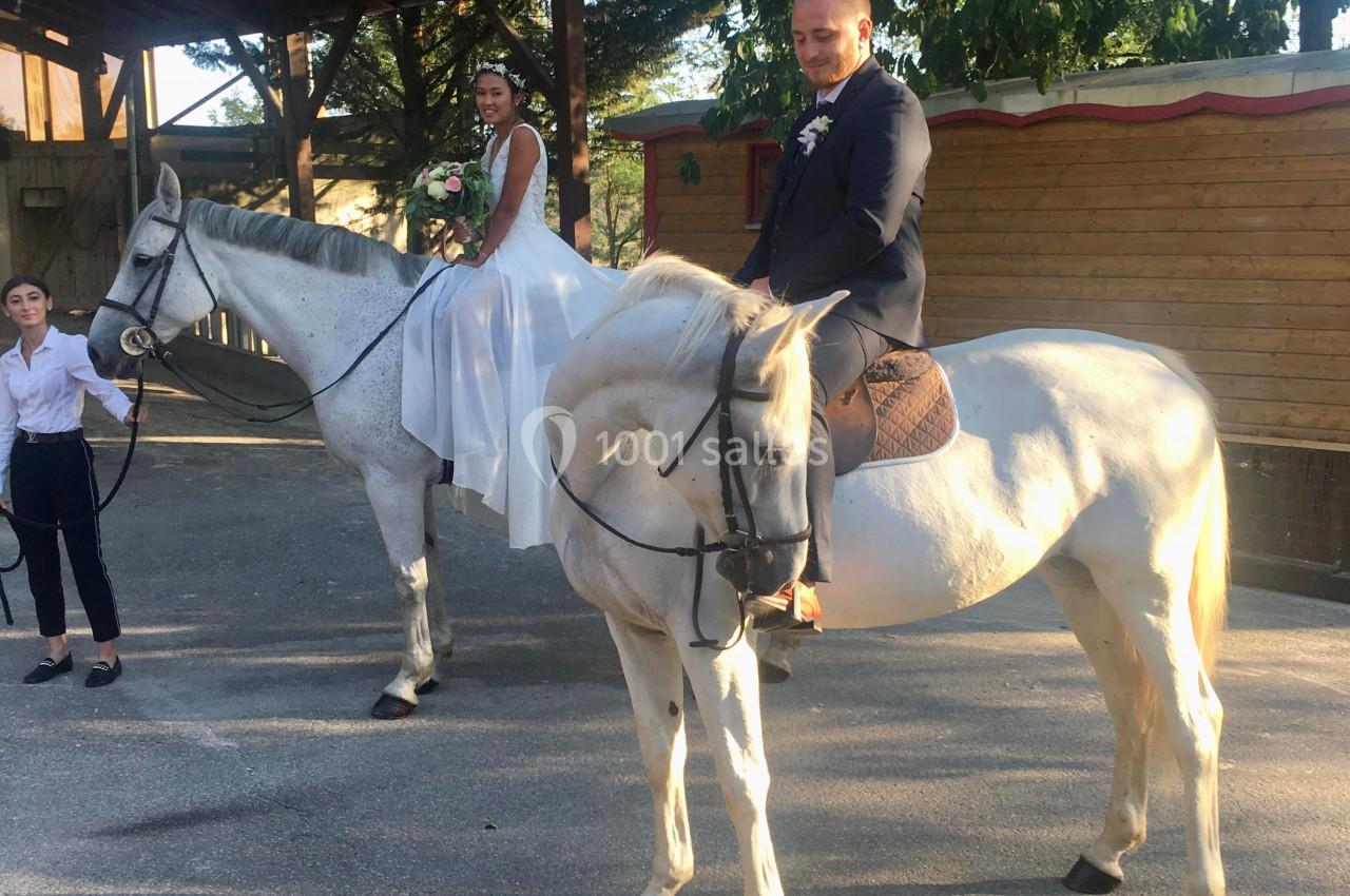 Un couple en tenue de mariage est assis sur deux chevaux blancs, accompagné d'une personne tenant un cheval.