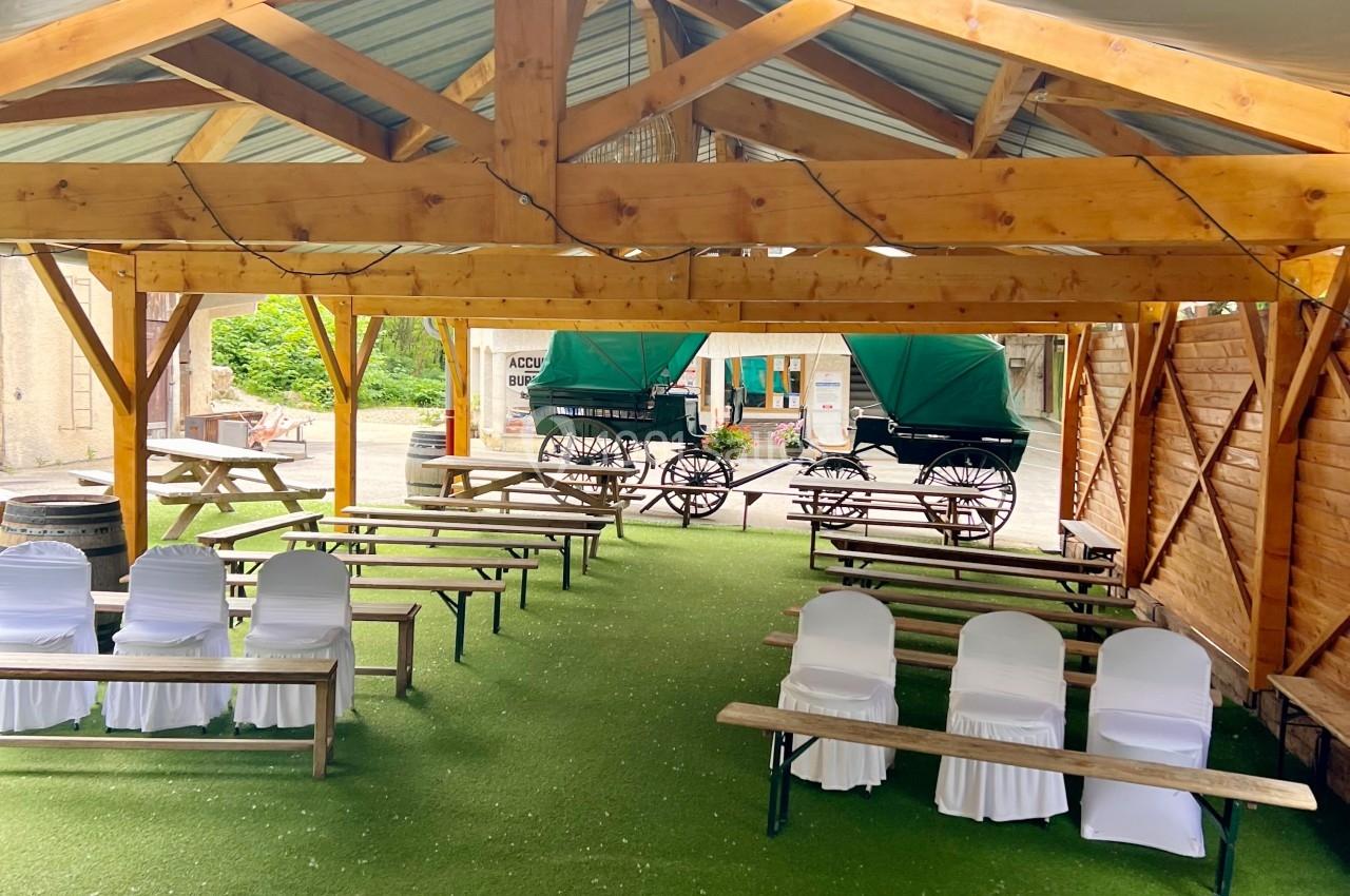 Structure en bois abritant des bancs et des chaises blanches sur un sol en gazon synthétique, avec des calèches en arrière…