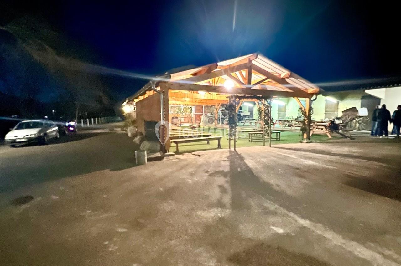 Abri en bois éclairé la nuit, entouré de bancs et situé sur un parking avec quelques voitures visibles.