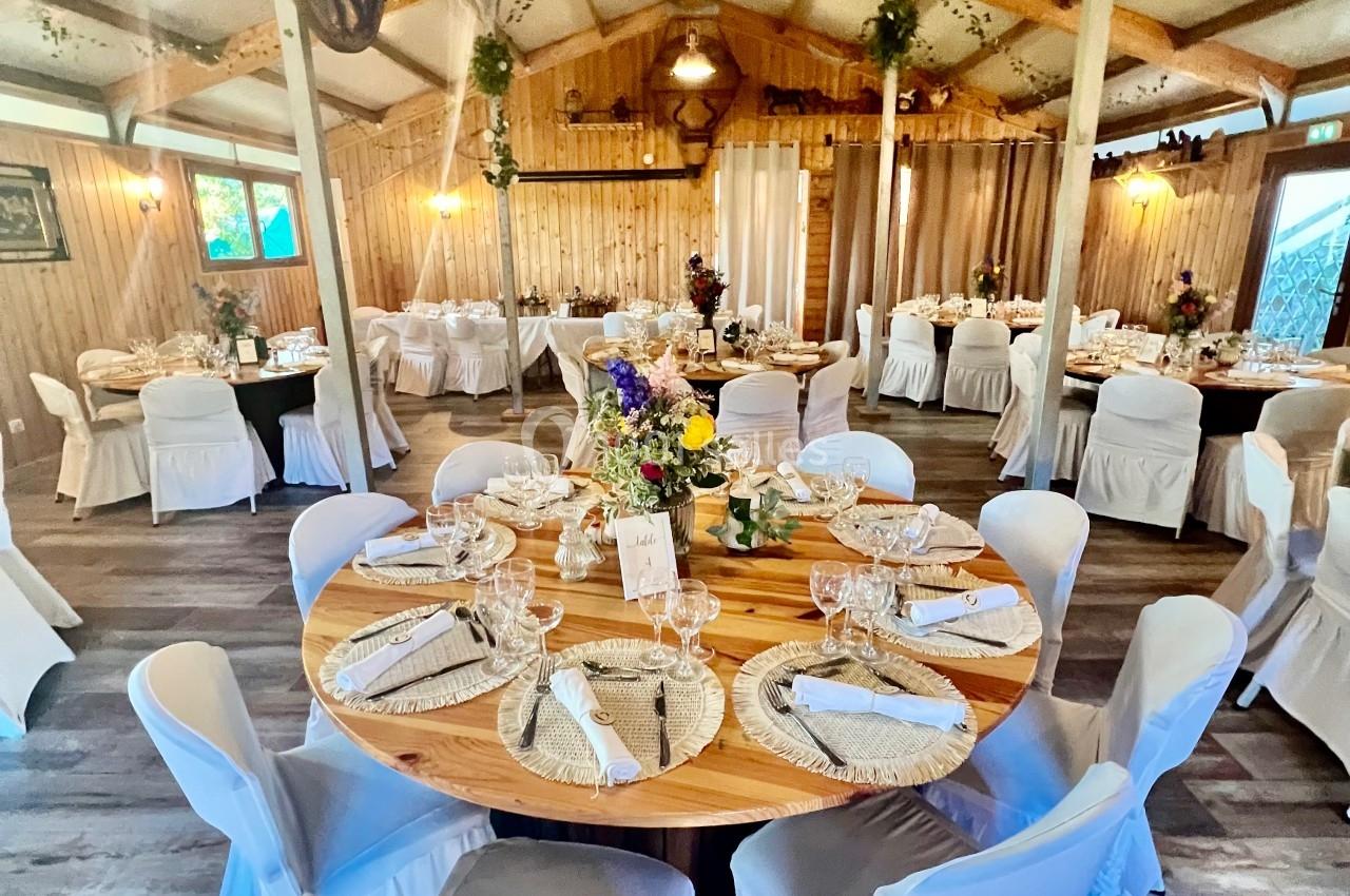 Salle de réception rustique décorée avec des tables rondes dressées, des chaises blanches et des éléments végétaux suspendus.