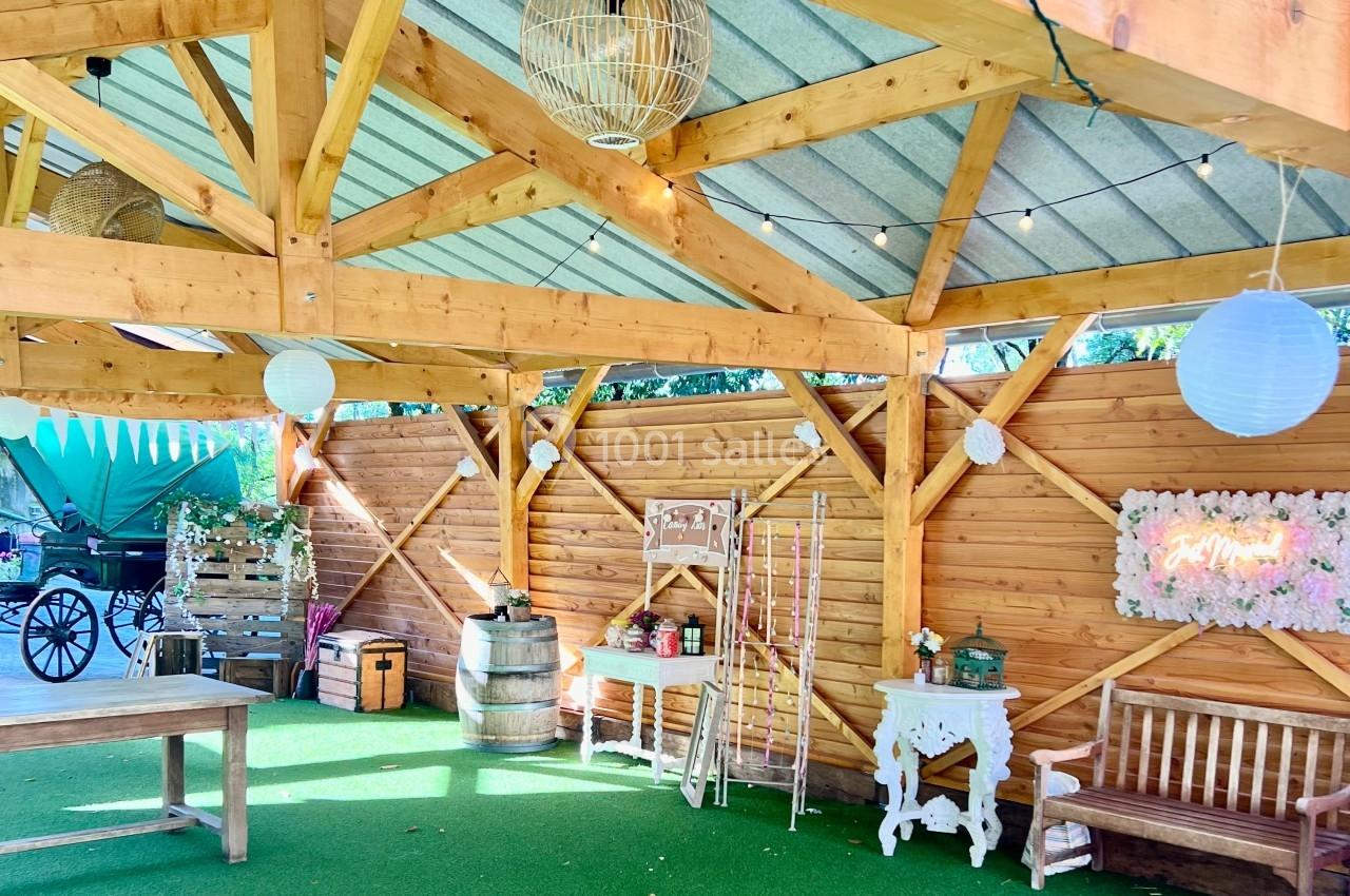 Structure en bois ouverte avec toit métallique, décorée de guirlandes lumineuses, mobilier rustique et fleurs.