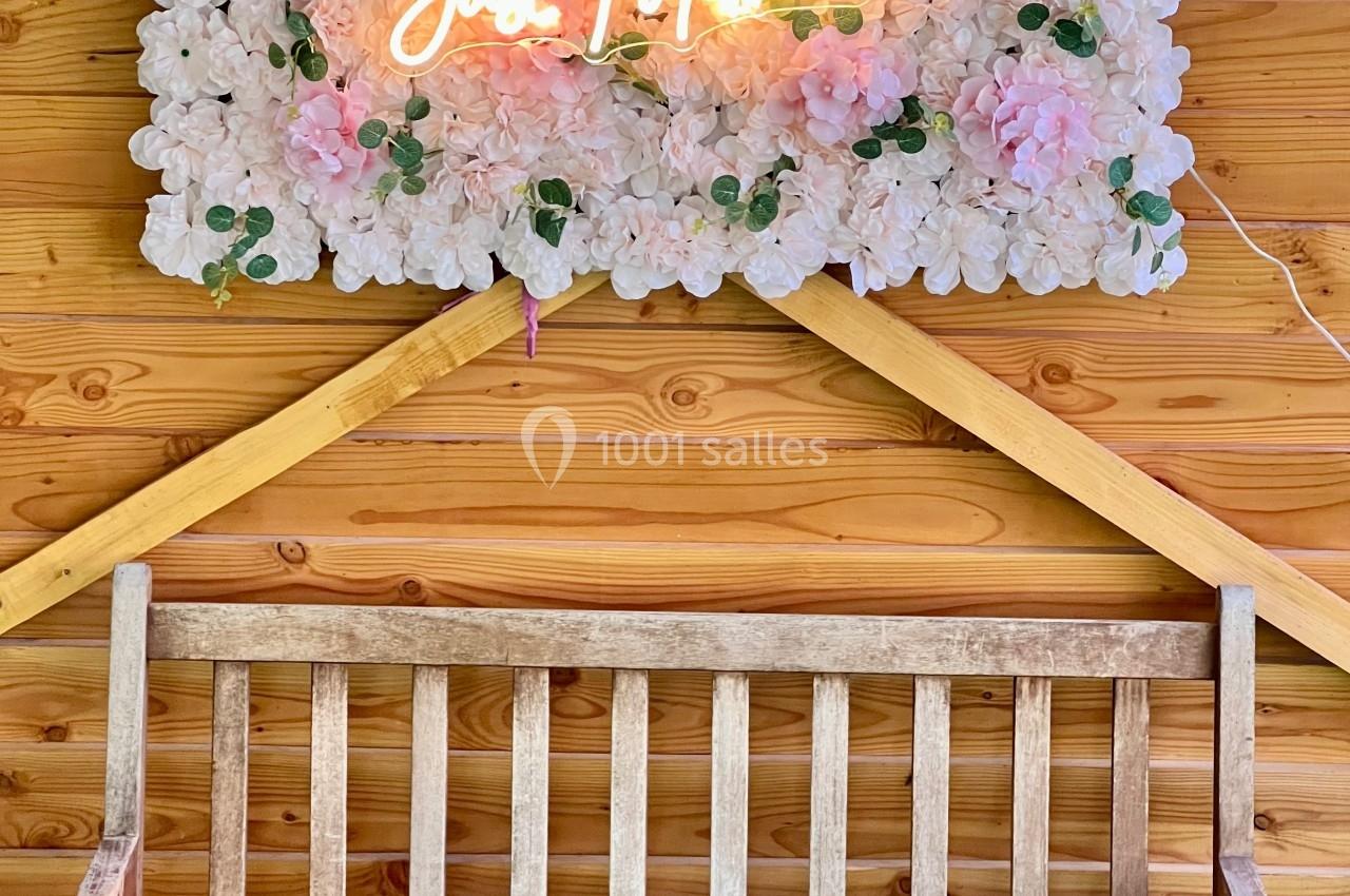 Banc en bois devant un panneau floral avec une inscription lumineuse ’Just Married’ sur un mur en bois.