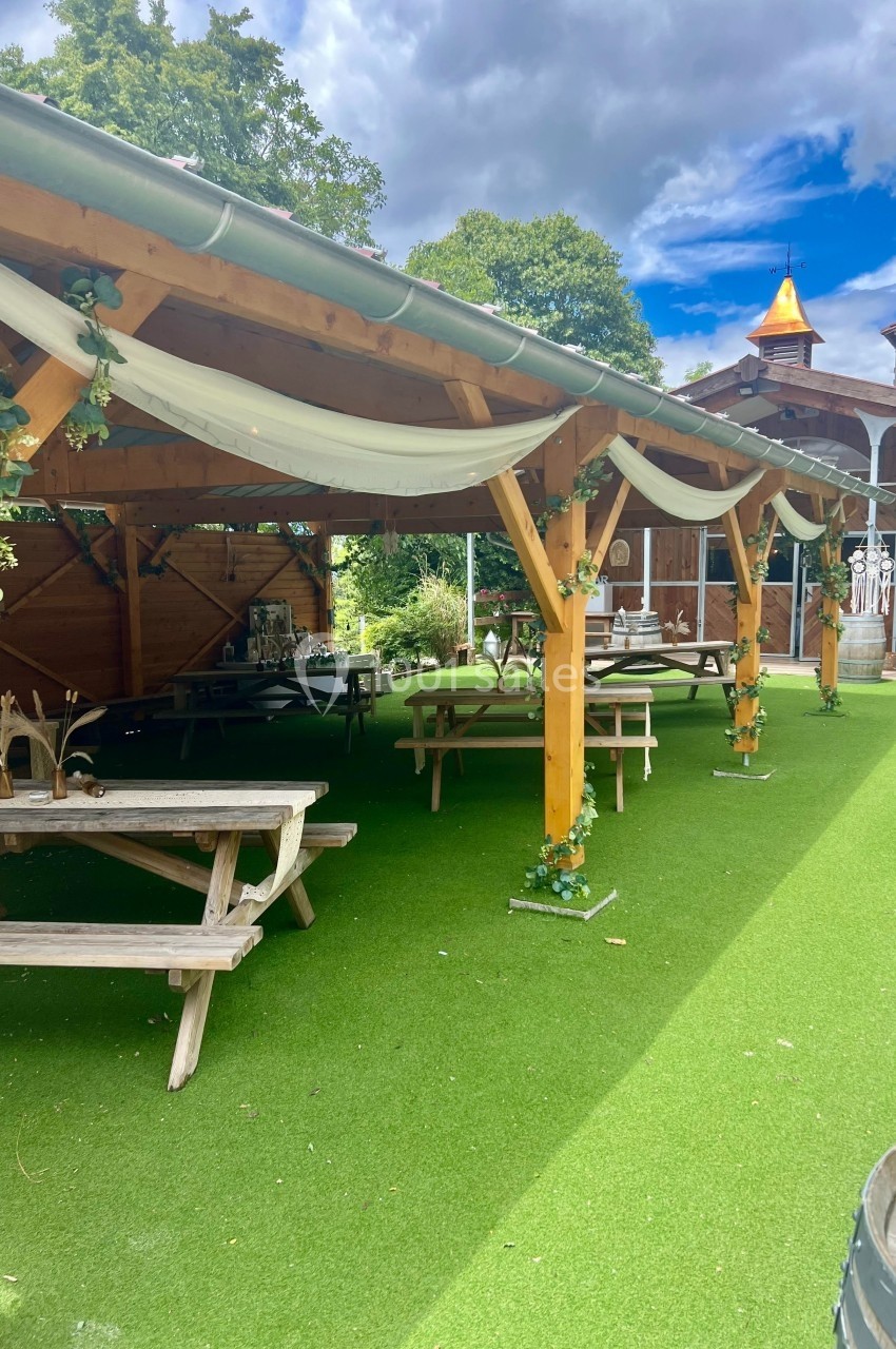 Abri en bois décoré de guirlandes et nappes blanches, avec tables de pique-nique sur une pelouse synthétique.