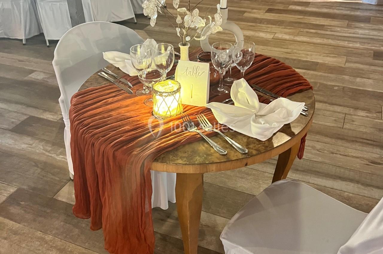 Table ronde décorée pour un dîner, avec nappe orange, vaisselle élégante, bougie allumée et centre de table floral.