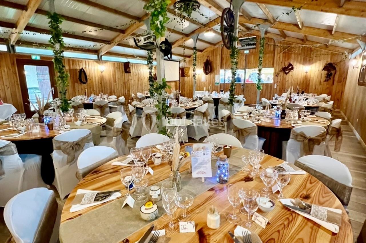Salle de réception décorée avec des tables rondes en bois, nappes blanches et éléments végétaux suspendus.
