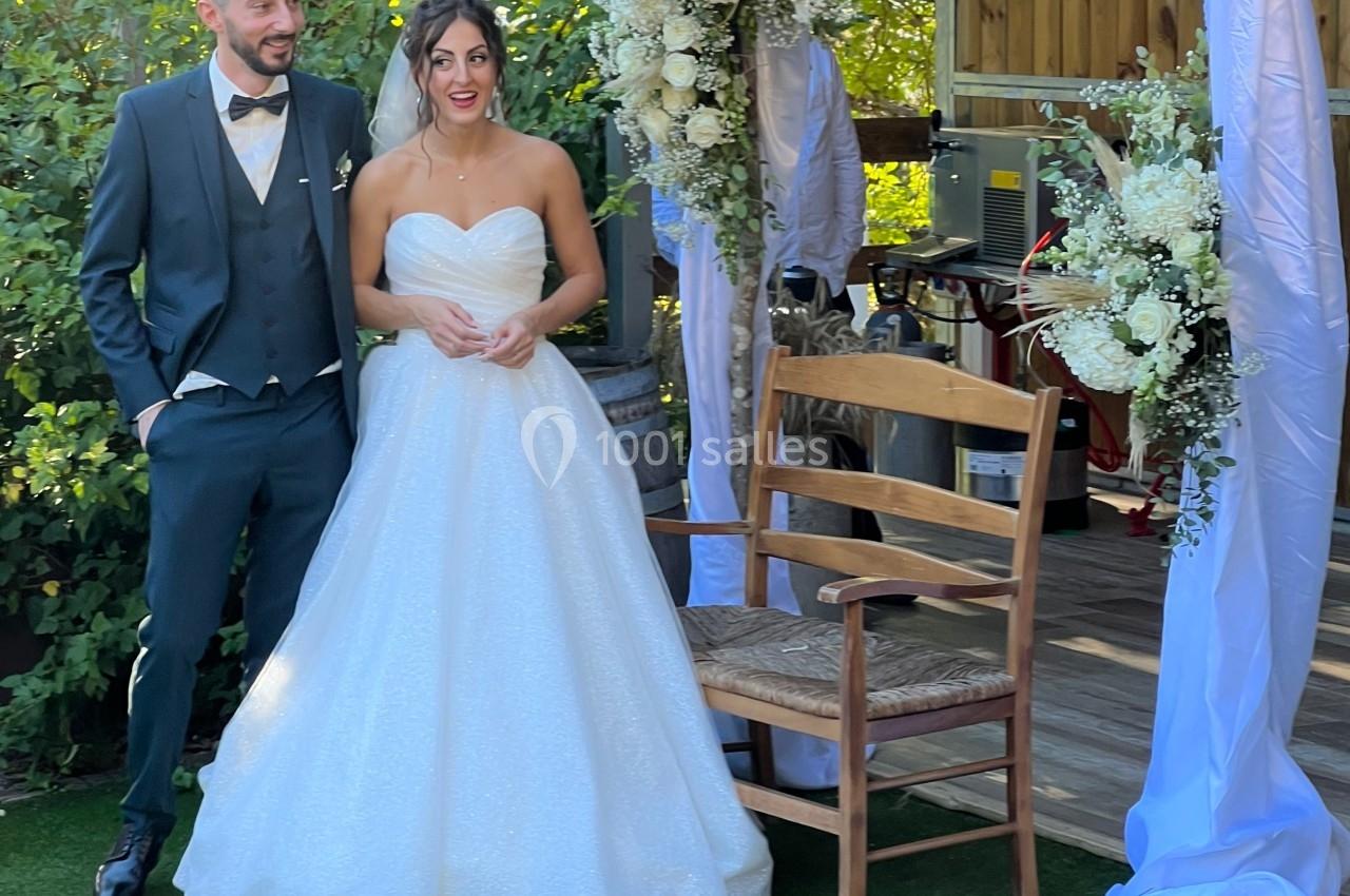 Un couple en tenue de mariage pose près d'une arche décorée de fleurs et de voiles dans un jardin.