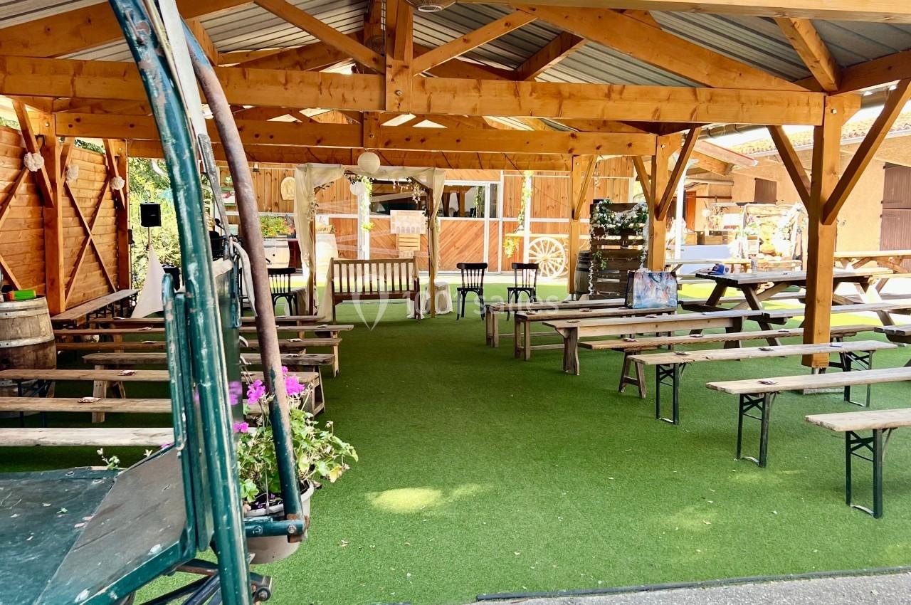 Structure en bois ouverte avec tables et bancs sur un sol en gazon synthétique, dans un espace extérieur aménagé.