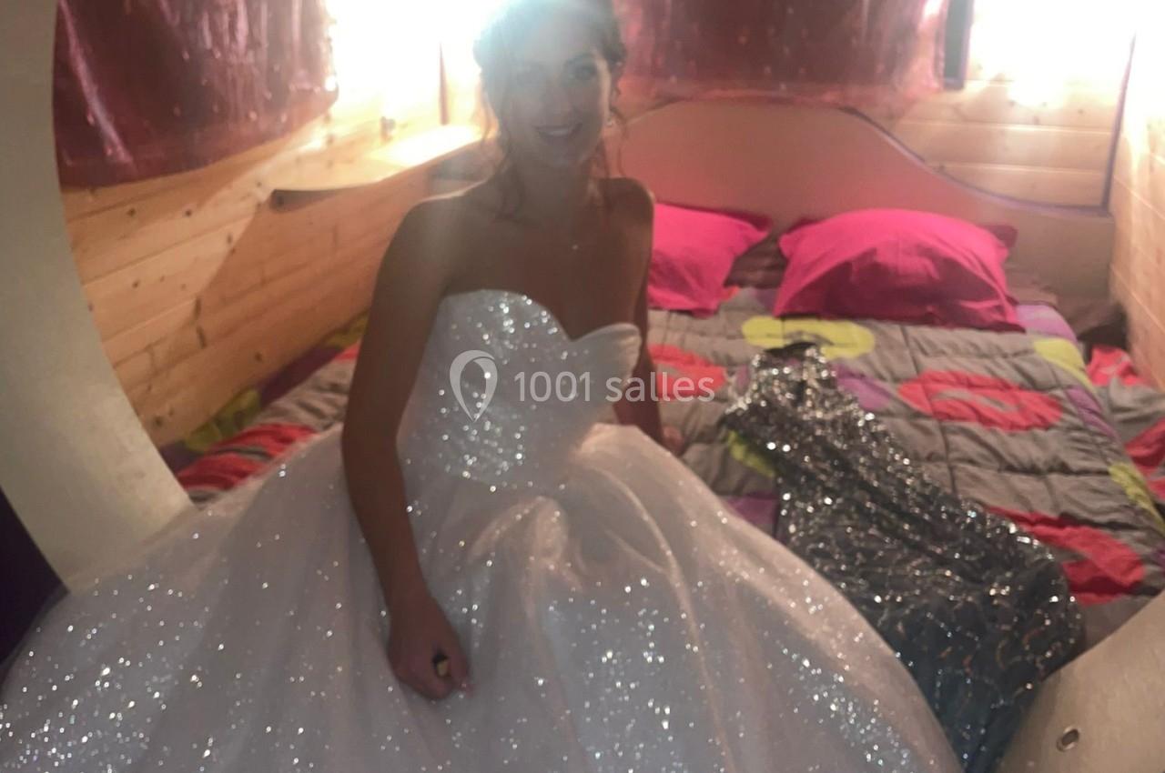 Femme en robe de mariée blanche scintillante assise dans une chambre en bois avec un lit coloré en arrière-plan.