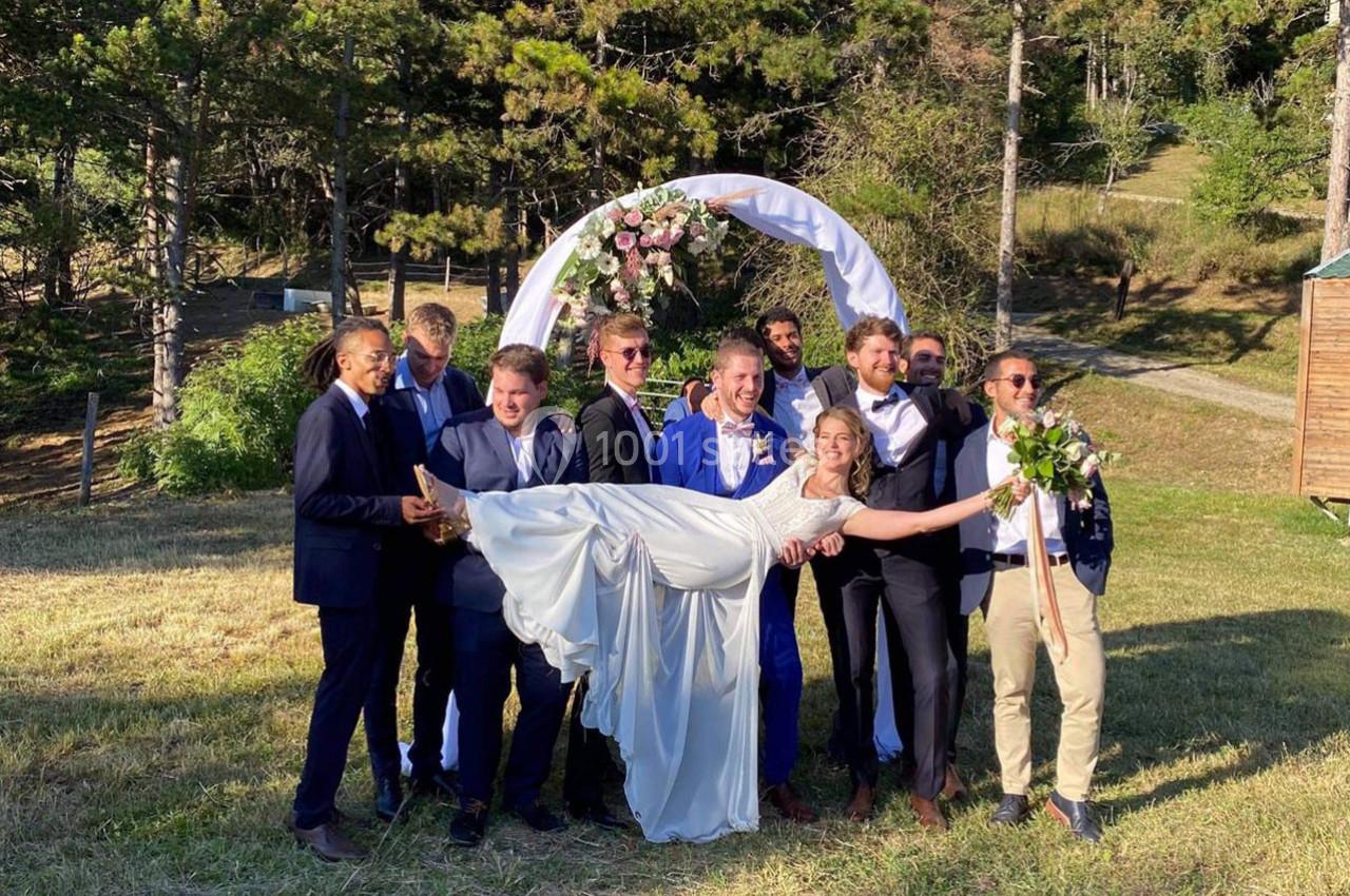 Un groupe d'hommes en costume porte une mariée en robe blanche devant une arche décorée de fleurs dans un jardin.