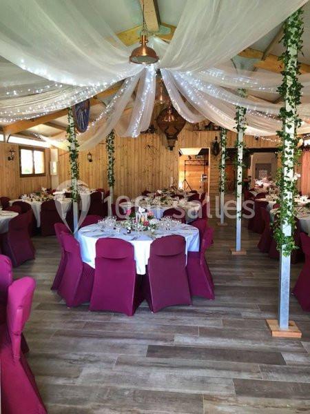 Salle de réception décorée avec des tables rondes, nappes blanches, chaises bordeaux et guirlandes lumineuses suspendues.