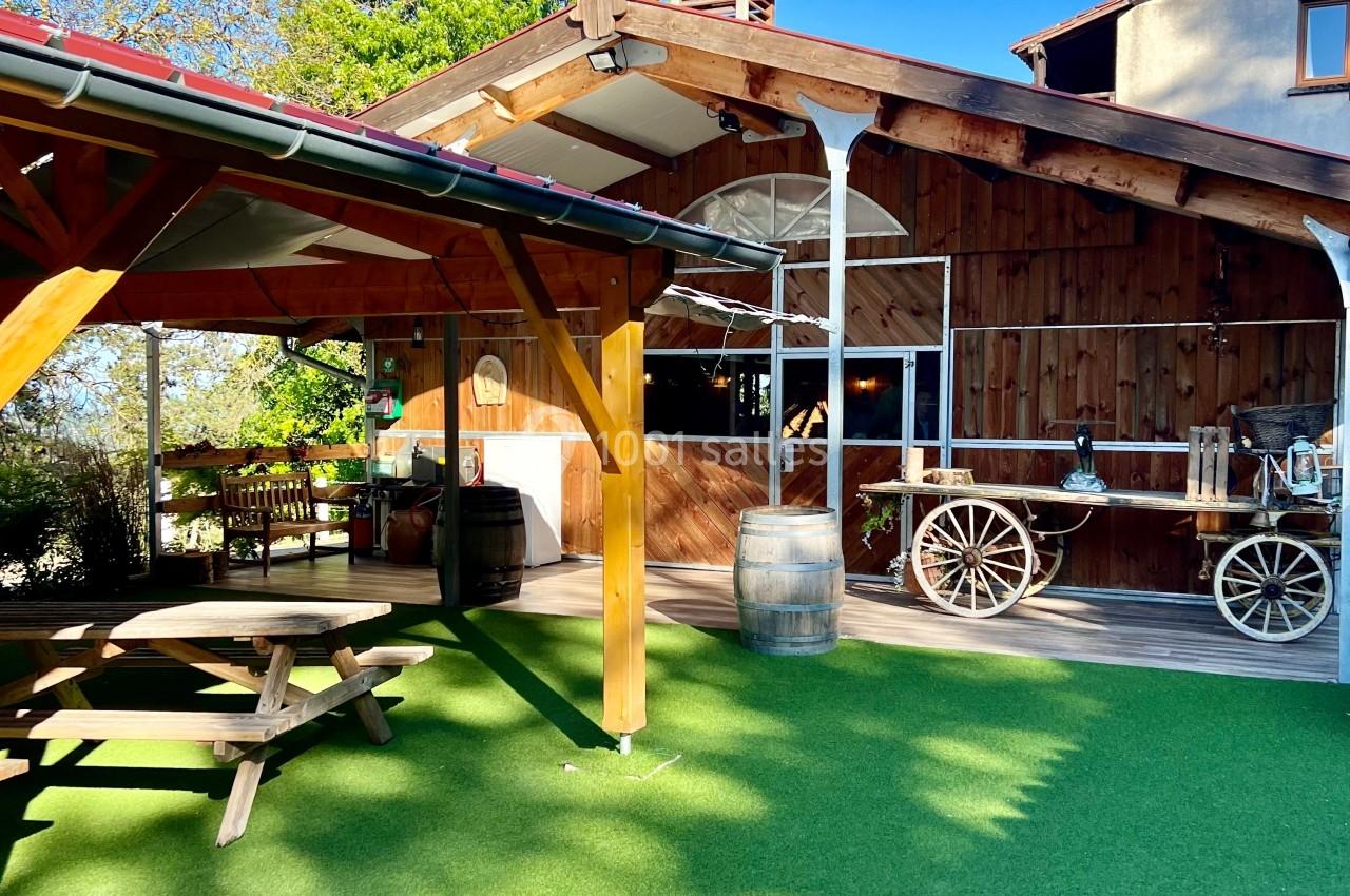 Cour extérieure avec gazon synthétique, tables en bois, charrette décorative et bâtiment en bois sous un ciel bleu.