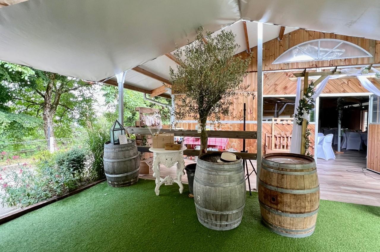 Terrasse couverte avec sol en gazon synthétique, tonneaux en bois, table blanche et arbre décoratif.