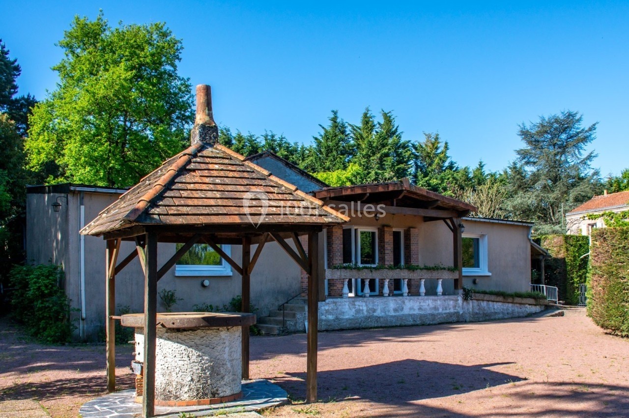 Maison avec façade beige, entourée de végétation, et un puits en pierre avec un toit en bois au premier plan.