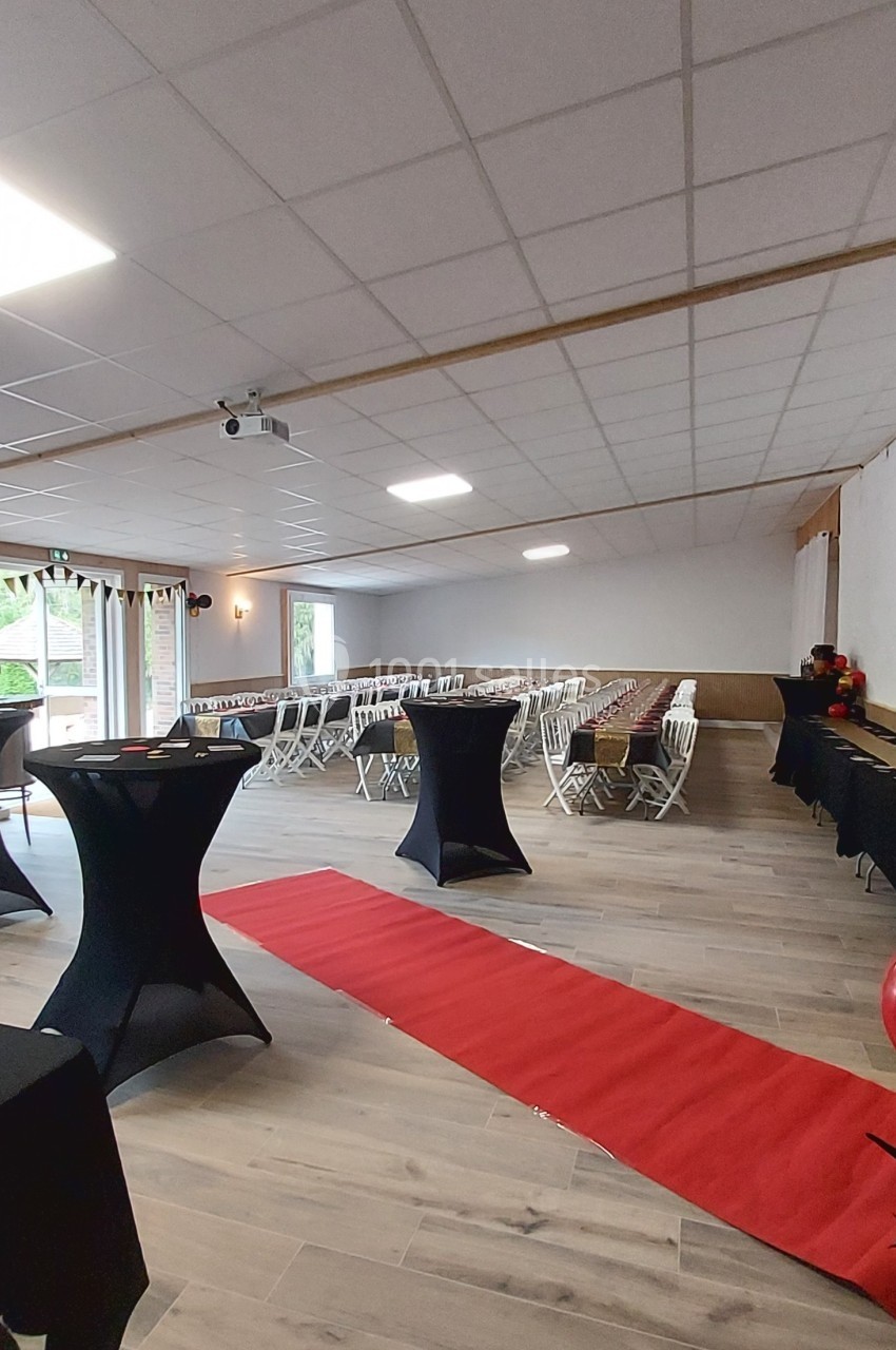 Salle de réception avec tables hautes, chaises alignées, tapis rouge et grandes fenêtres laissant entrer la lumière…