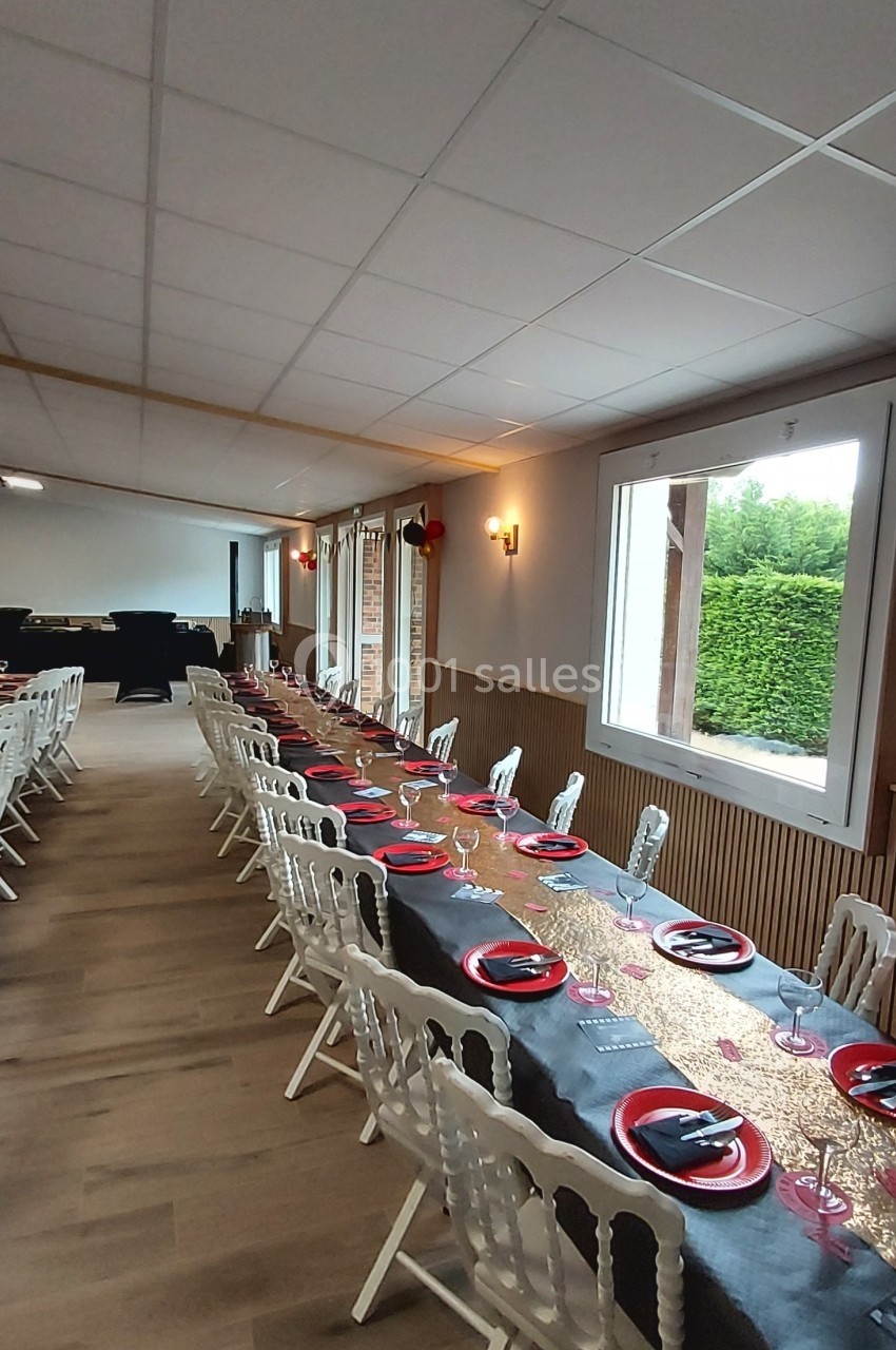 Salle décorée avec une longue table dressée, nappes noires et dorées, assiettes rouges, chaises blanches, grandes fenêtres.