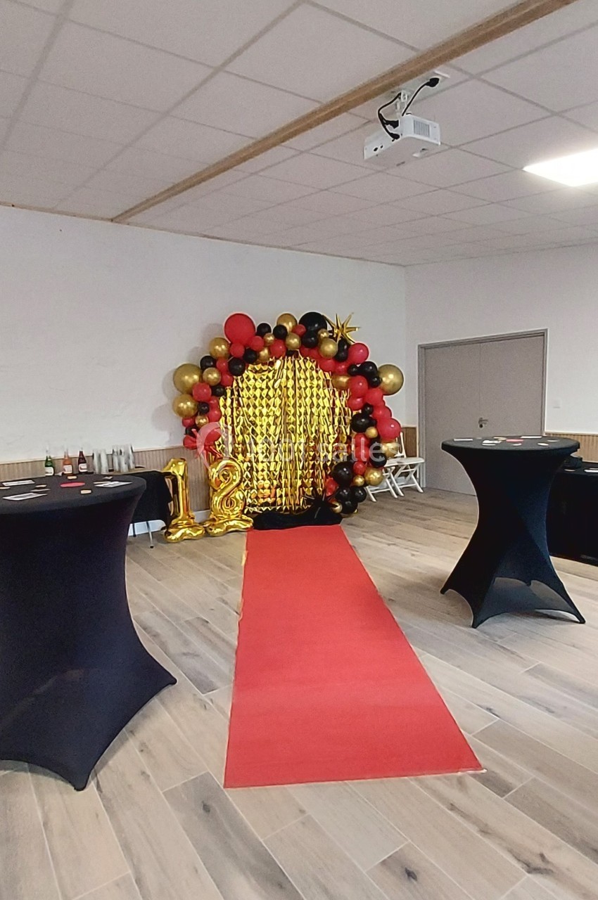 Décoration festive avec arche de ballons rouges, noirs et dorés, rideau scintillant et tapis rouge dans une salle.