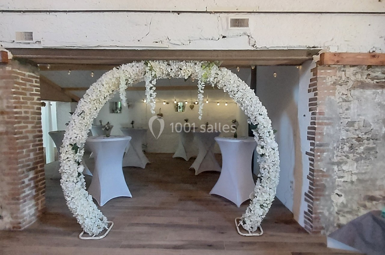 Arche florale blanche décorant l'entrée d'une salle en pierre avec des tables hautes recouvertes de nappes blanches.