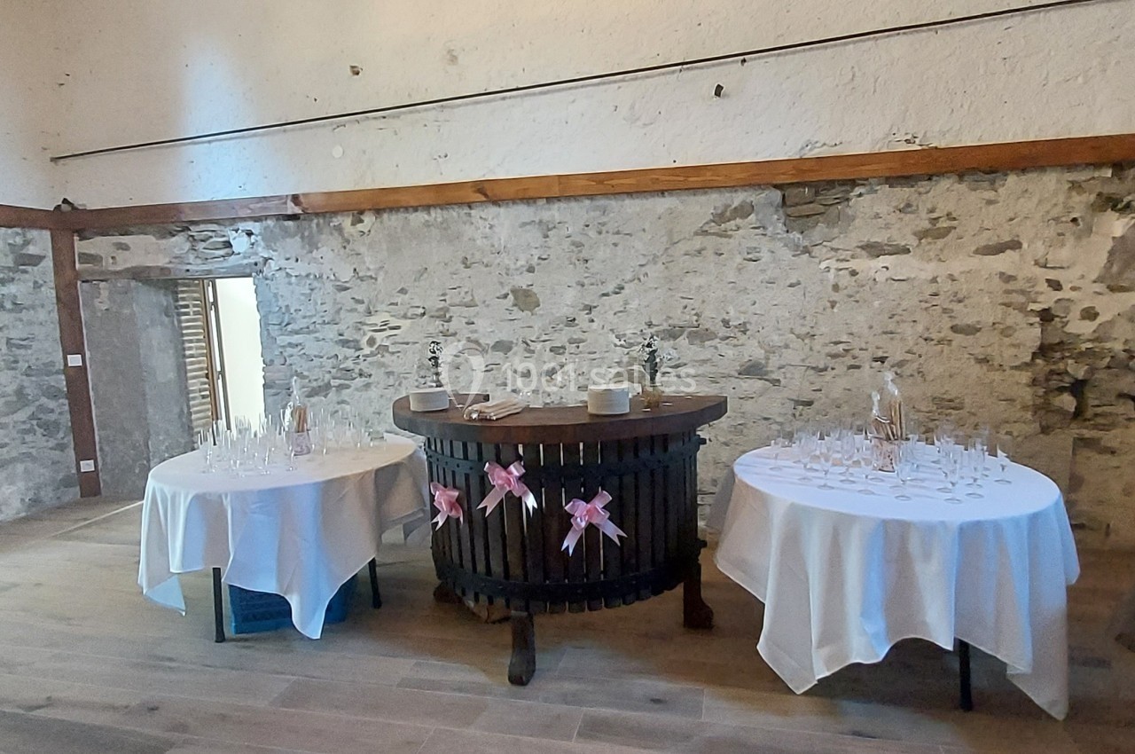 Salle en pierre avec deux tables rondes dressées pour un événement, décorées de nappes blanches et rubans roses.