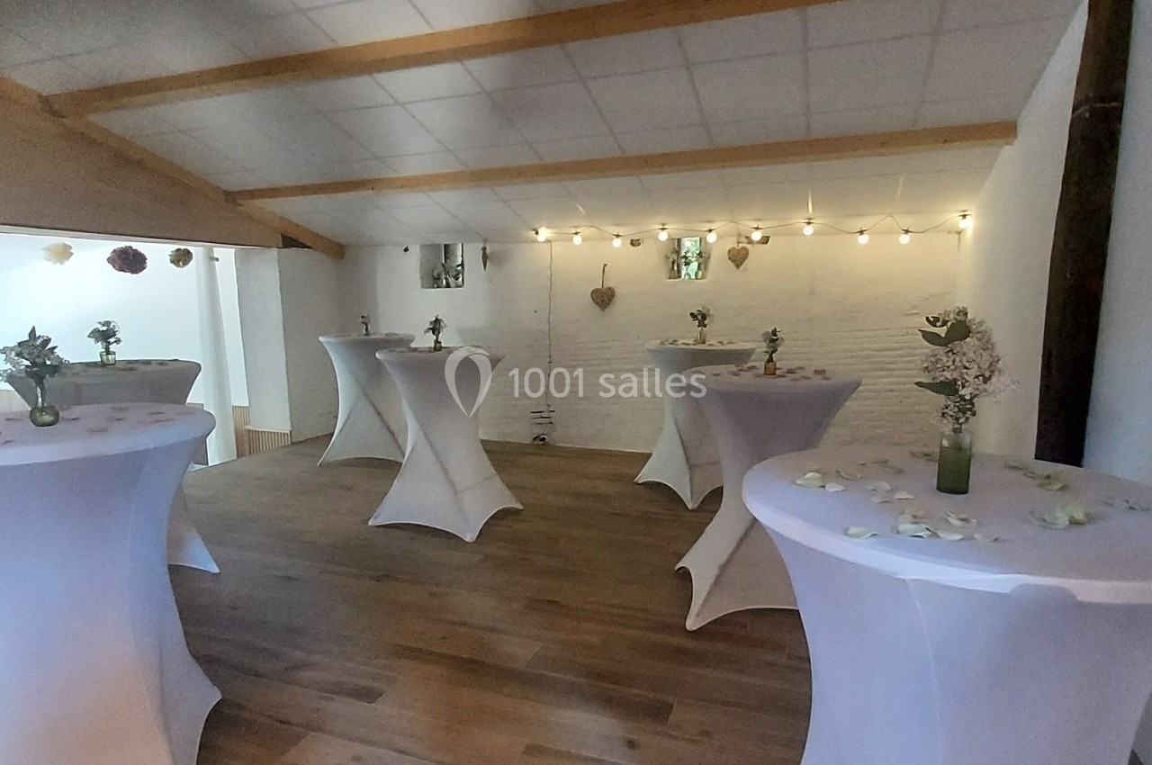 Salle décorée avec des tables hautes recouvertes de nappes blanches, ornées de fleurs et de pétales, éclairée par des…
