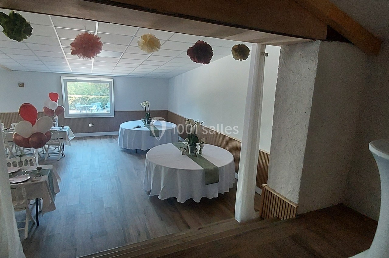 Salle décorée pour un événement, avec tables rondes dressées, ballons et guirlandes suspendues.