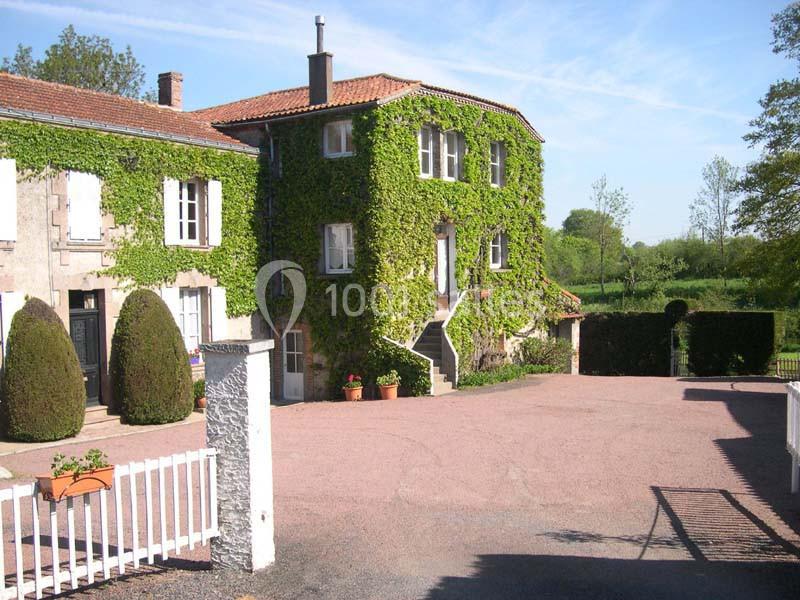 Maison en pierre recouverte de lierre, avec une cour pavée et un portail blanc, entourée de verdure.