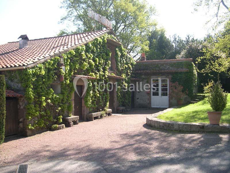 Maison en pierre avec façade recouverte de vigne, toit en tuiles rouges et allée pavée entourée de verdure.