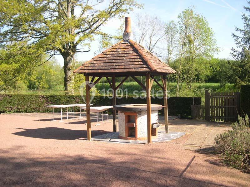Abri en bois avec toit en tuiles protégeant un barbecue en pierre, situé dans une cour extérieure arborée.