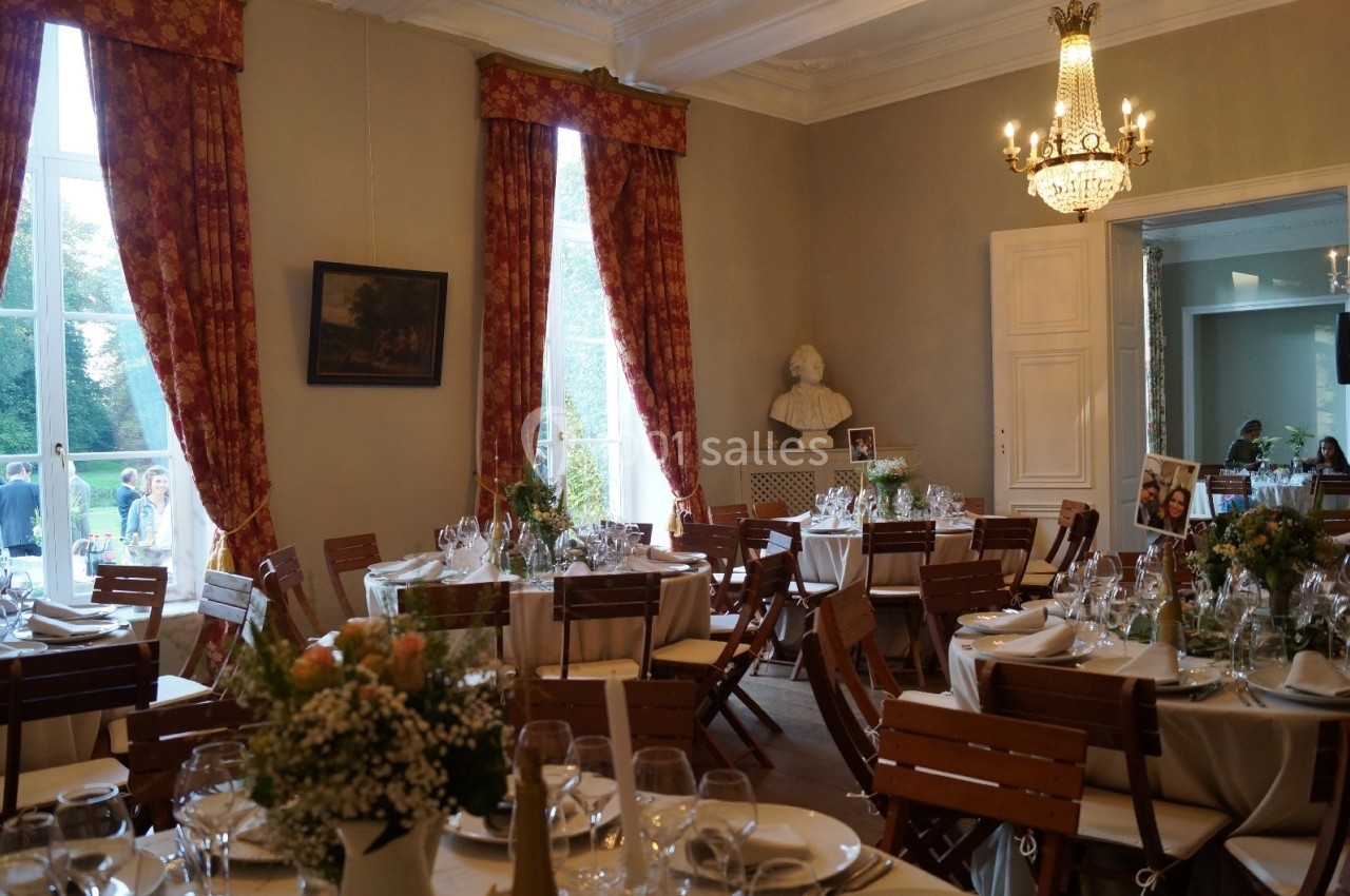 Salle élégante avec tables dressées pour un événement, décorée de rideaux rouges, lustre et statue en arrière-plan.