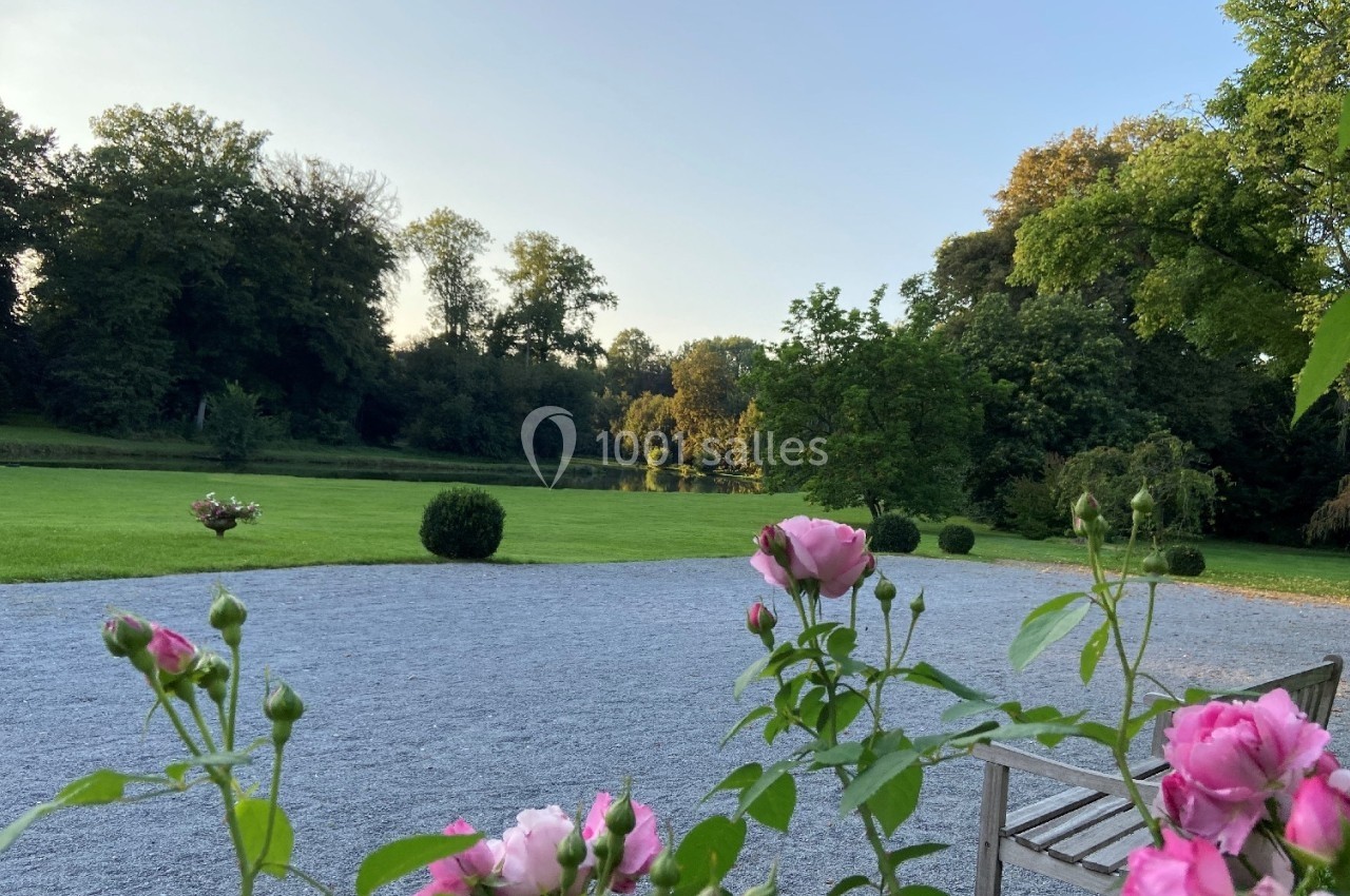 Allée gravillonnée bordée de roses avec vue sur une pelouse, des arbres et un plan d'eau sous un ciel dégagé.