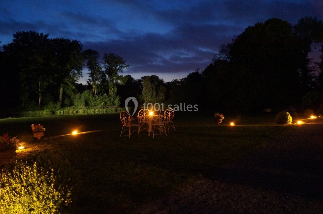 Chaises et table en fer forgé éclairées par des lumières au sol dans un jardin au crépuscule.