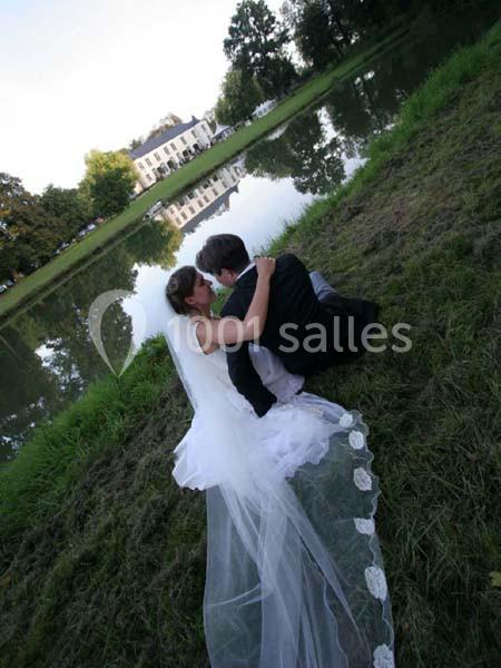 Un couple de mariés assis sur l'herbe près d'un lac, avec un bâtiment blanc reflété dans l'eau.