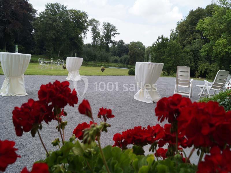 Espace extérieur avec tables hautes drapées, chaises blanches et géraniums rouges au premier plan, entouré de verdure.