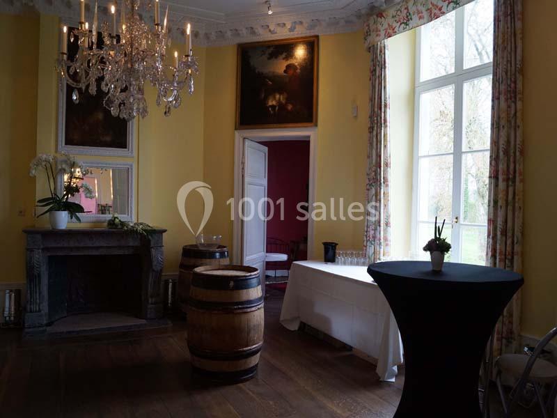 Salle élégante avec murs jaunes, cheminée, lustre, tonneaux en bois, table drapée et grande fenêtre donnant sur un jardin.