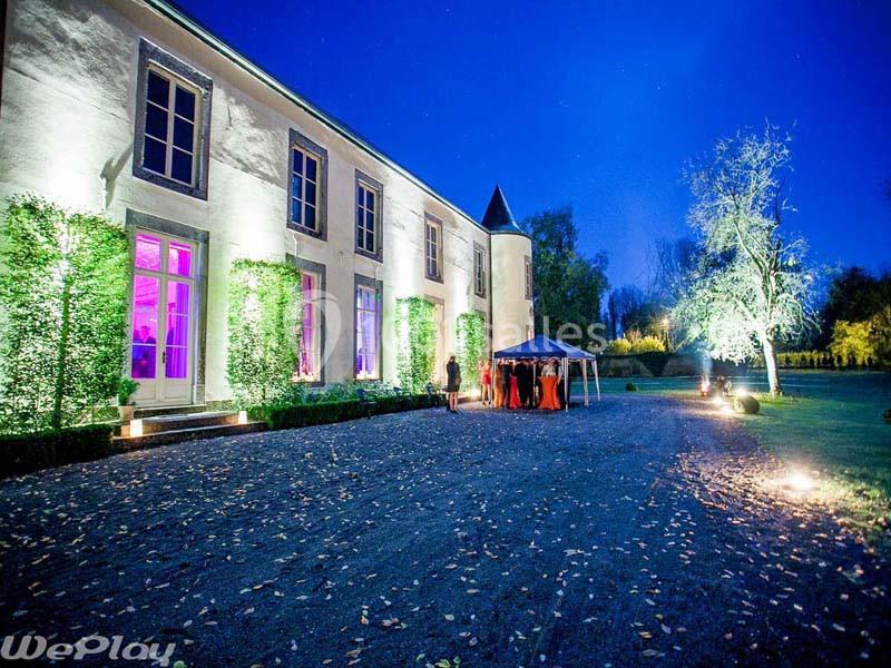 Façade éclairée d'un château entouré de végétation, avec des invités rassemblés sous un chapiteau le soir.