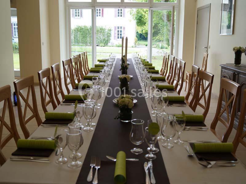 Table dressée avec des couverts, serviettes vertes et décoration florale, dans une salle lumineuse avec vue sur un jardin.