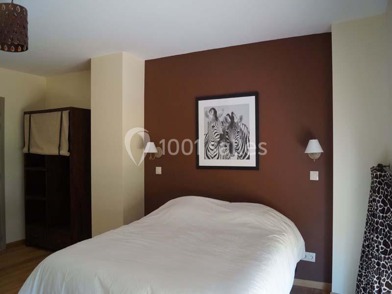 Chambre avec un lit double, mur brun décoré d'un tableau de zèbres, deux appliques murales et une armoire sombre.