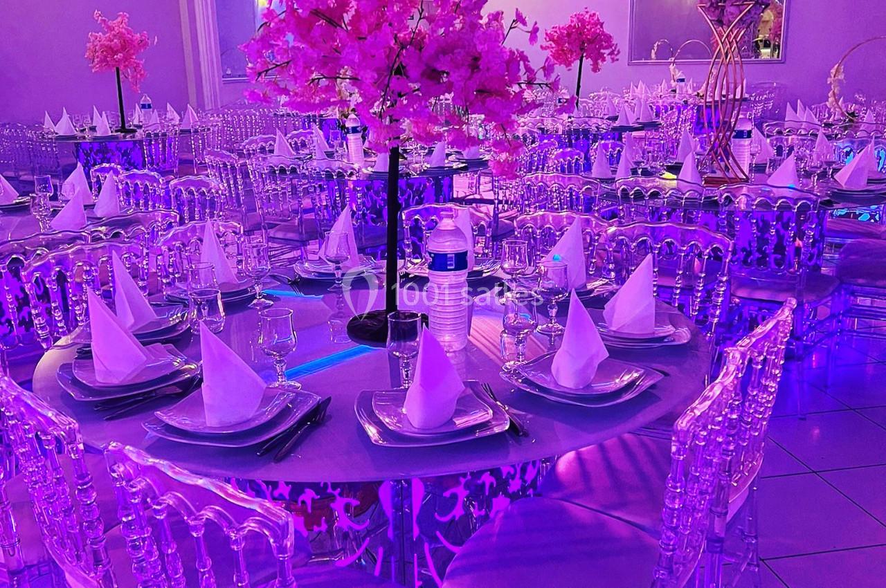 Salle de réception décorée avec des tables rondes, nappes violettes, serviettes pliées et centre de table floral rose.