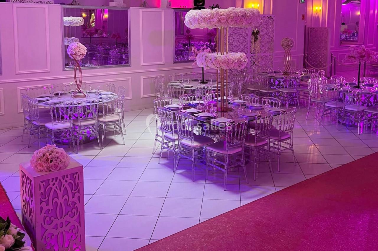 Salle de réception décorée avec des tables rondes, chaises transparentes et arrangements floraux sous un éclairage rose.
