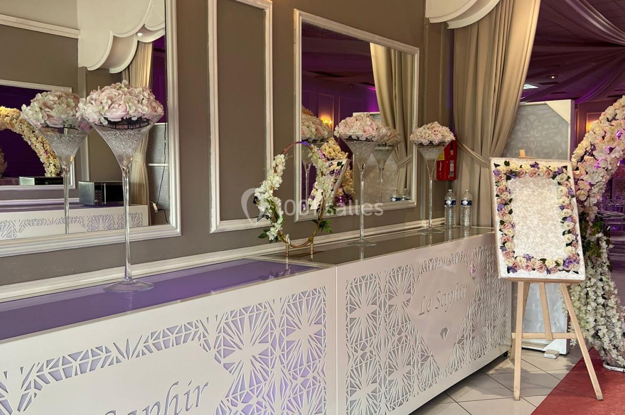 Comptoir décoré avec des fleurs blanches et roses, miroir en arrière-plan, rideaux et panneau floral à droite.