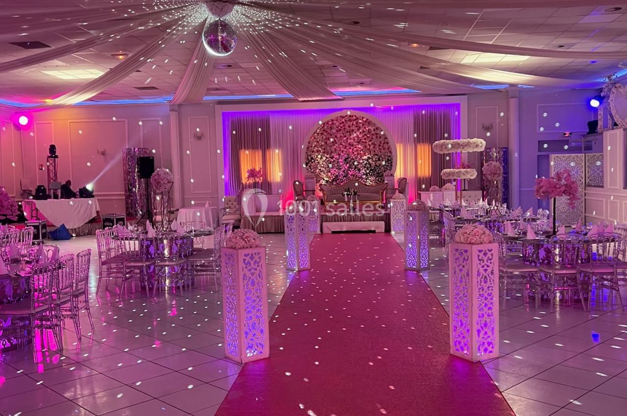 Salle de réception décorée avec un tapis rouge, des lumières colorées, des tables dressées et une scène ornée de fleurs.