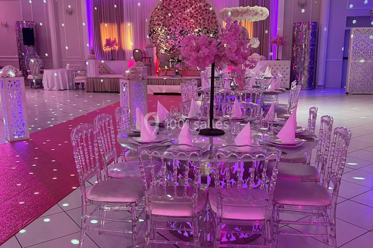 Salle de réception décorée avec des tables rondes, chaises transparentes et éclairages colorés, ambiance festive.