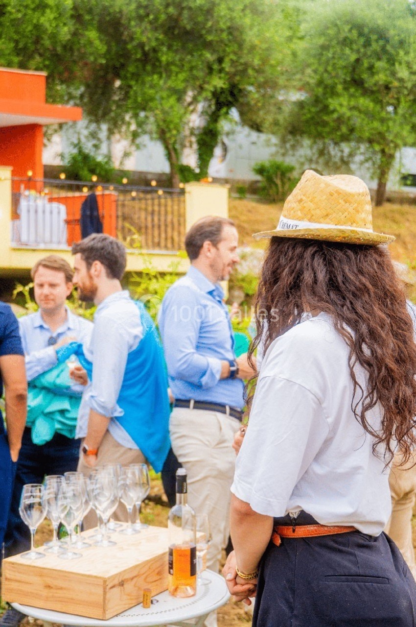 Une femme portant un chapeau observe un groupe de personnes discutant près d'une table avec des verres et une bouteille.