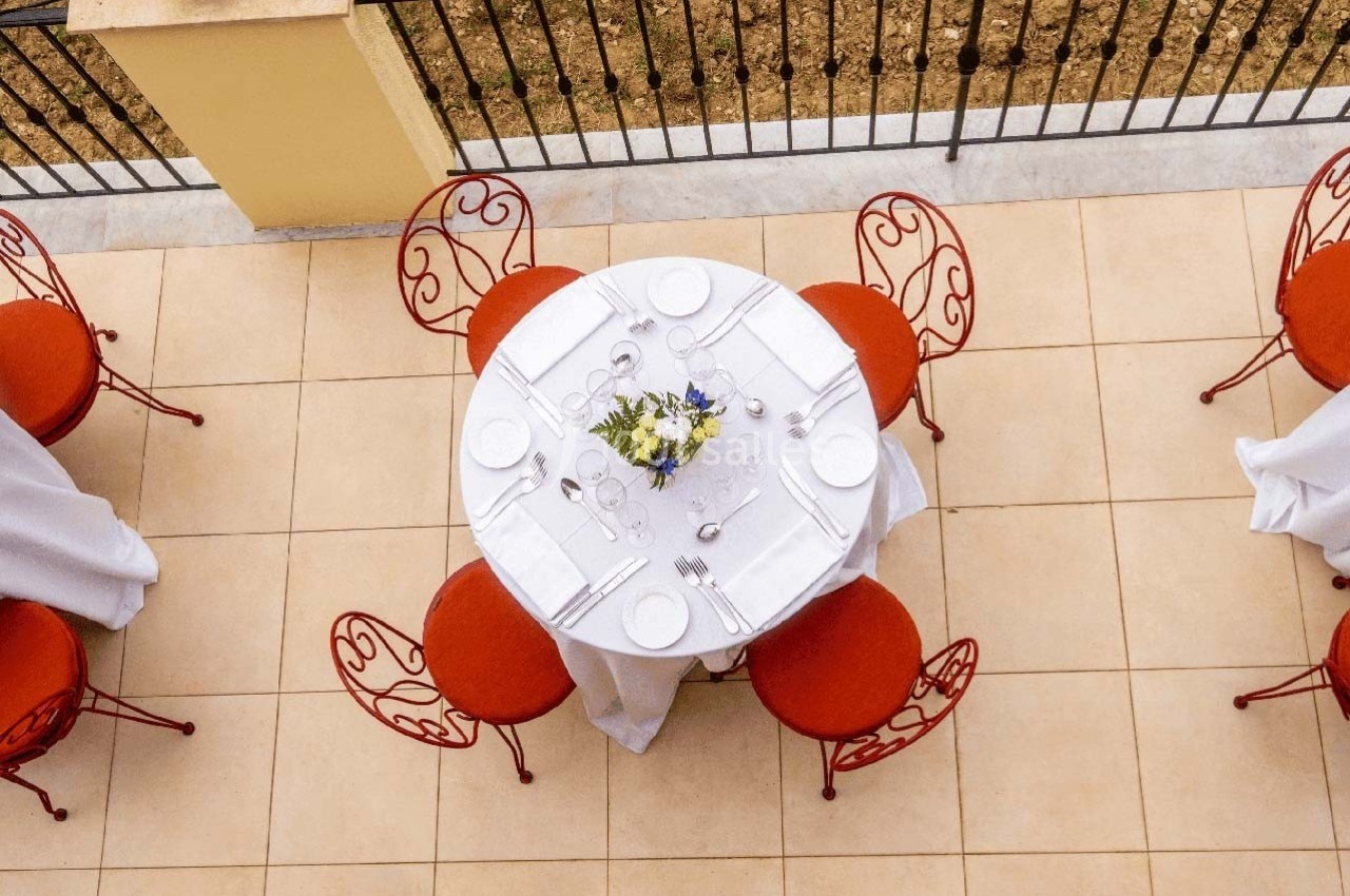 Table ronde dressée avec vaisselle blanche et bouquet de fleurs, entourée de chaises rouges sur une terrasse carrelée.