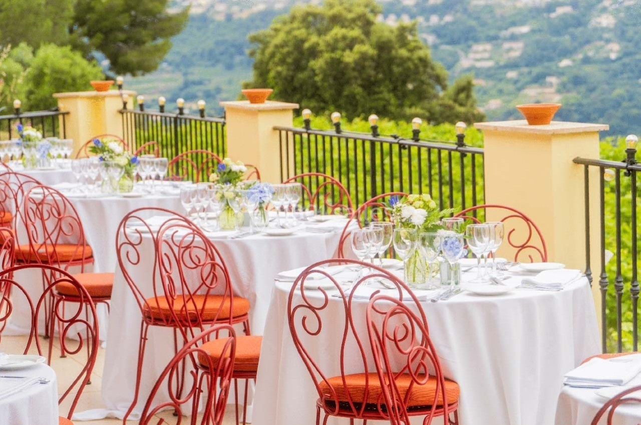 Tables dressées avec nappes blanches et chaises rouges sur une terrasse surplombant un paysage verdoyant.