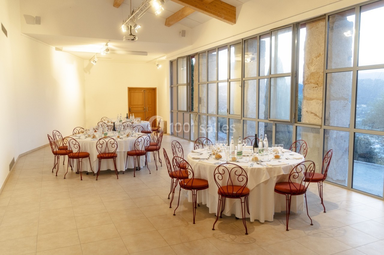 Salle lumineuse avec tables rondes dressées, chaises en fer forgé et grandes baies vitrées donnant sur l'extérieur.