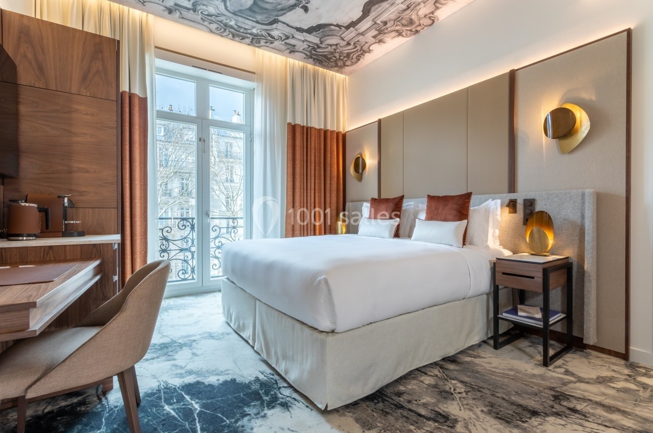 Chambre d'hôtel lumineuse avec lit double, bureau en bois, fauteuil, et grande fenêtre donnant sur un balcon.