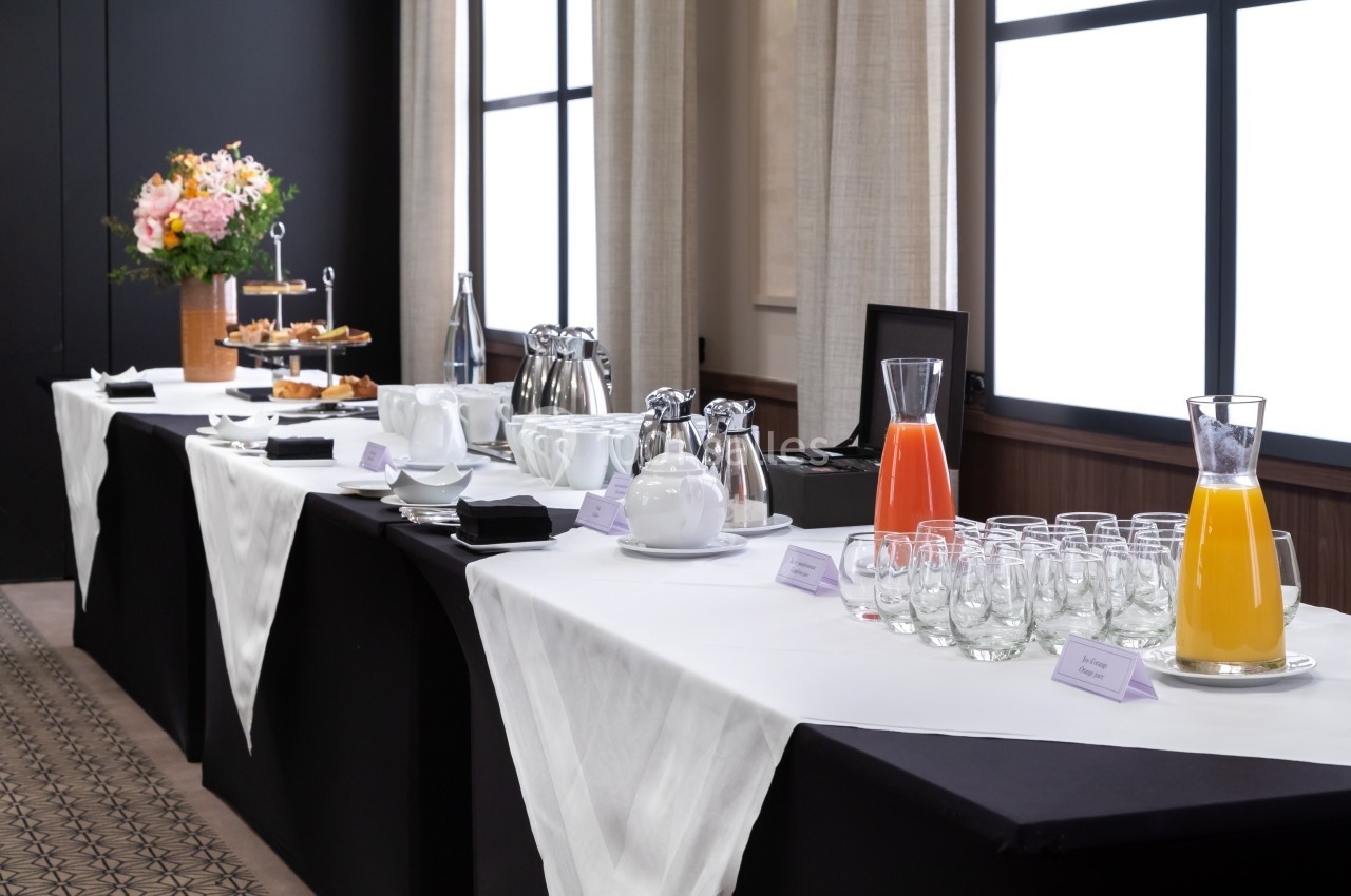 Table de buffet avec carafes de jus, tasses, assiettes, viennoiseries et un bouquet de fleurs en arrière-plan.